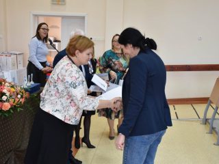 Kobieta w kwiecistej marynarce wręcza dokument uczestniczce w granatowej marynarce.