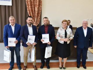 Trzej mężczyźni z dyplomami i torbami upominkowymi pozują obok organizatorów uroczystości.