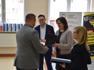 Mężczyzna w szarej marynarce odbiera nagrodę od organizatorów stojących przy stole.