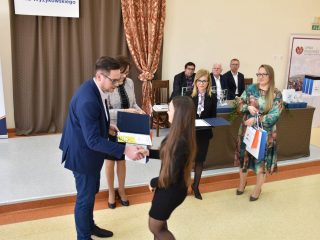 Mężczyzna w niebieskim garniturze wręcza otwarte etui młodej kobiecie w czarnej sukience.
