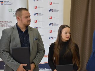 Mężczyzna w szarej marynarce i młoda kobieta trzymają czarne teczki podczas uroczystości nagród.