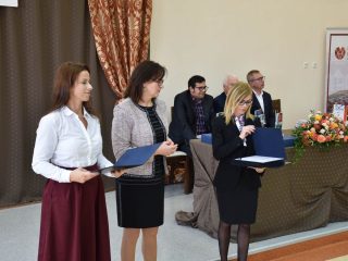 Trzy organizatorki stoją z otwartymi teczkami; kobieta w okularach odczytuje tekst do mikrofonu.