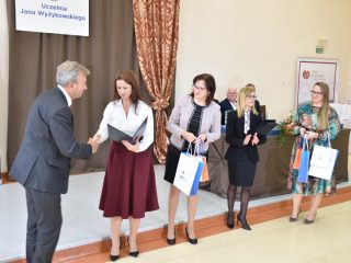 Siwowłosy mężczyzna odbiera gratulacje od kobiety w białej bluzce i bordowej spódnicy.