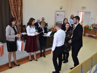 Grupa uczestników stoi przed organizatorkami odczytującymi dokumenty podczas wręczania nagród.