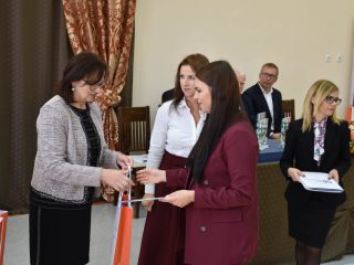 Kobieta w bordowej marynarce odbiera torbę upominkową od organizatorki w szarej marynarce.