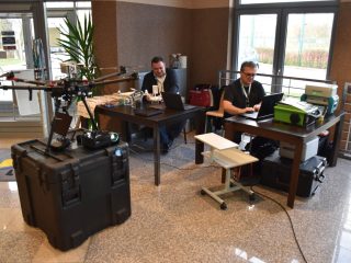 Stanowisko pokazowe z dronami, aparaturą i komputerami w holu podczas konferencji naukowej.