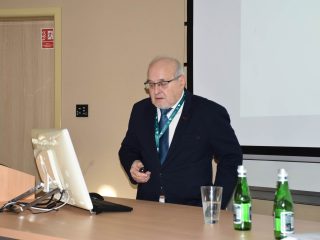 Prelegent w okularach przemawia przy komputerze w sali konferencyjnej.