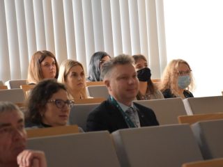 Uczestnicy konferencji naukowej słuchają wykładu w sali z szarymi fotelami.