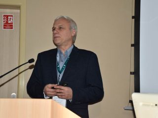 Siwowłosy prelegent z identyfikatorem przemawia przy mównicy.
