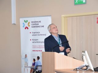 Prelegent spogląda na ekran, stojąc przy mównicy i banerze Regionalnego Centrum Konferencyjnego.