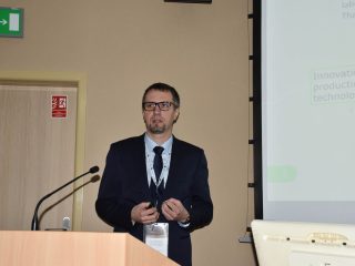 Mężczyzna w okularach prowadzi wystąpienie przy mównicy, trzymając pilot do prezentacji.