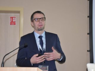 Prelegent gestykuluje obiema dłońmi podczas wystąpienia na konferencji naukowej.