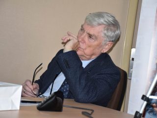 Siwowłosy uczestnik konferencji siedzi przy stole, trzymając okulary i słuchając wystąpienia.