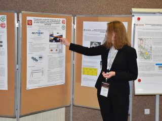 Kobieta w czarnej marynarce wskazuje schemat urządzenia na posterze naukowym.