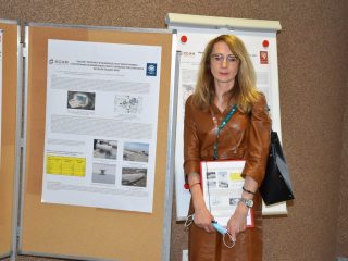 Kobieta w okularach i brązowej sukience trzyma dokumenty, pozując przy posterze naukowym.