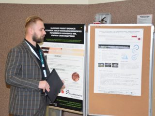 Mężczyzna w marynarce w kratę trzyma czarną teczkę, stojąc obok posterów naukowych.