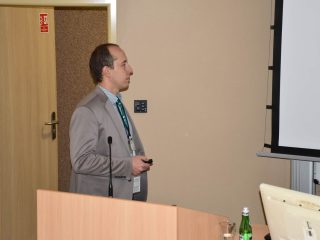 Prelegent w szarej marynarce z pilotem prezentacyjnym przy mównicy.