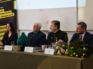 Uczestnicy uroczystości odznaczeń CBJ siedzą przy stole, uśmiechając się i spoglądając w lewo.