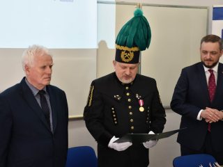 Mężczyzna w mundurze górniczym, czaku z zielonym pióropuszem i białych rękawiczkach odczytuje dokument z otwartej teczki.