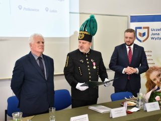 Mężczyzna w mundurze górniczym trzyma otwartą teczkę i zwraca się do stojącego obok siwowłosego mężczyzny.