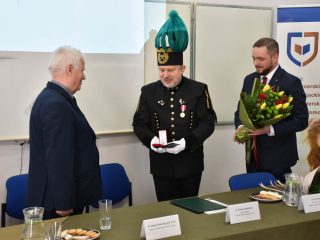 Przedstawiciel w mundurze górniczym prezentuje otwarte czerwone etui z odznaczeniem; obok stoi mężczyzna z bukietem.