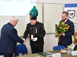 Siwowłosy mężczyzna ściska dłoń przedstawiciela w mundurze górniczym trzymającego etui z odznaczeniem.