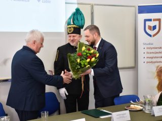 Mężczyzna w garniturze wręcza bukiet siwowłosemu uczestnikowi uroczystości, a obok stoi przedstawiciel w mundurze górniczym.
