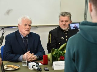 Siwowłosy mężczyzna wypowiada się przy stole przed mikrofonami i kamerą; obok siedzi przedstawiciel w mundurze górniczym.