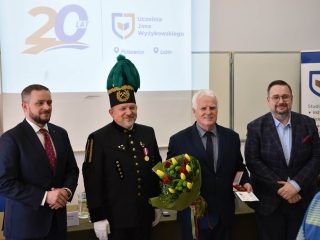 Czterej mężczyźni pozują do zdjęcia podczas uroczystości odznaczeń CBJ, z bukietem kwiatów i etui z medalem.