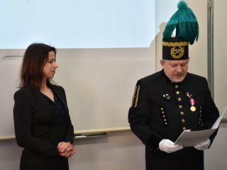 Mężczyzna w mundurze górniczym i czaku z zielonym pióropuszem czyta dokument obok kobiety w czarnej marynarce.