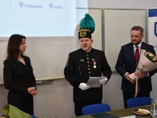 Przedstawiciel w mundurze górniczym odczytuje dokument; obok stoją kobieta i mężczyzna z bukietem.