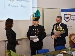 Kobieta słucha przedstawiciela w mundurze górniczym trzymającego dokument, a mężczyzna obok trzyma bukiet.