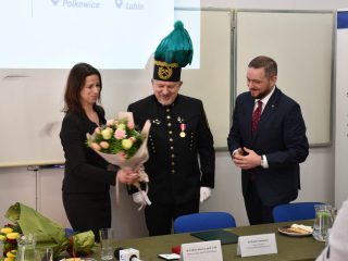 Kobieta trzyma bukiet różowych kwiatów obok przedstawiciela w mundurze górniczym i mężczyzny w garniturze.