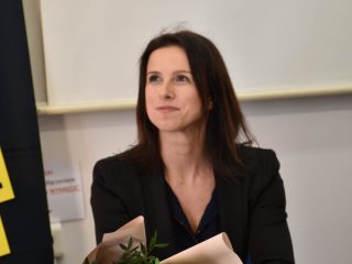Kobieta w czarnej marynarce siedzi przy stole z bukietem różowych kwiatów.