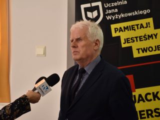 Siwowłosy mężczyzna udziela wywiadu przed banerem Uczelni Jana Wyżykowskiego.
