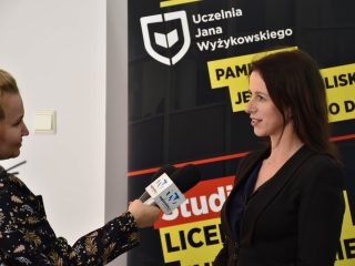 Kobieta udziela wywiadu reporterce trzymającej mikrofon przed banerem uczelni.