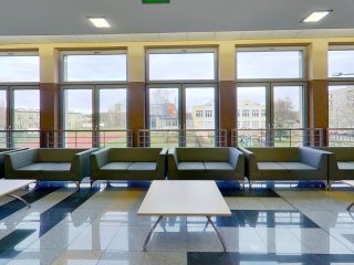 Rząd zielonych foteli i jasne stoliki przy dużych oknach w holu.