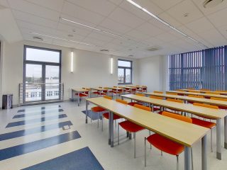 Jasna sala ćwiczeniowa z rzędami stolików i czerwono-pomarańczowymi krzesłami.