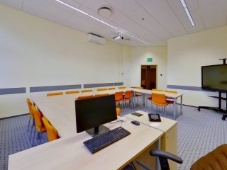 Sala UJW ze stołami ustawionymi w podkowę, komputerem prowadzącego i dużym monitorem.