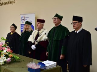 Przedstawiciele uczelni w togach stoją przy stole z kwiatami podczas zakończenia roku PUTW.