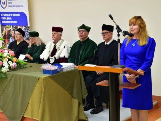 Kobieta w niebieskiej sukience przemawia przy mównicy obok przedstawicieli uczelni w togach.