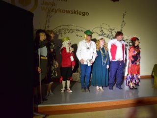 Zespół teatralny w kolorowych kostiumach stoi w rzędzie na scenie.