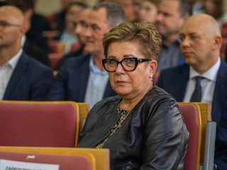 Kobieta w okularach i ciemnej kurtce siedzi wśród gości inauguracji.