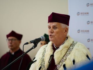 Rektor w uroczystej todze, birecie i łańcuchu przemawia do mikrofonu.