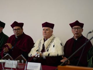 Przedstawiciele uczelni w togach siedzą przy stole prezydialnym, część z nich klaszcze.