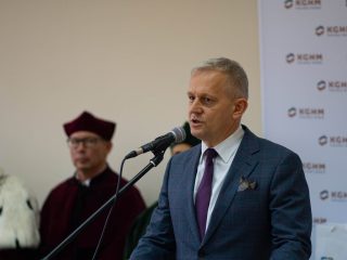 Siwowłosy mężczyzna w niebieskim garniturze wygłasza przemówienie do mikrofonu.