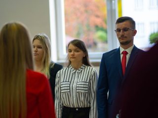 Dwie studentki i student w garniturze stoją przy sztandarze podczas inauguracji.