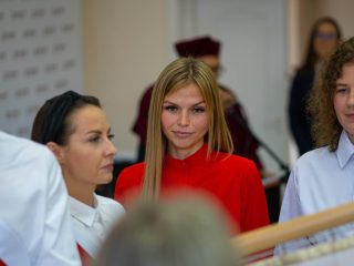 Studentka w czerwonej bluzce stoi przy sztandarze obok pozostałych uczestników ślubowania.