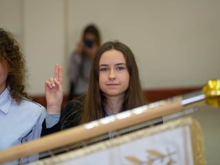 Studentka unosi prawą dłoń z dwoma palcami podczas ślubowania przy sztandarze.
