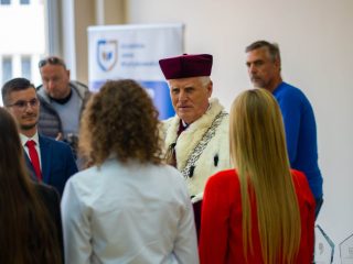 Rektor w uroczystej todze stoi przed grupą studentów przyjmowanych do społeczności akademickiej.